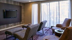 Una habitación de hotel moderna con un televisor de pantalla plana montado en la pared, un ventanal con cortinas traslúcidas, una alfombra estampada y una zona de estar con dos sofás y una mesita.
