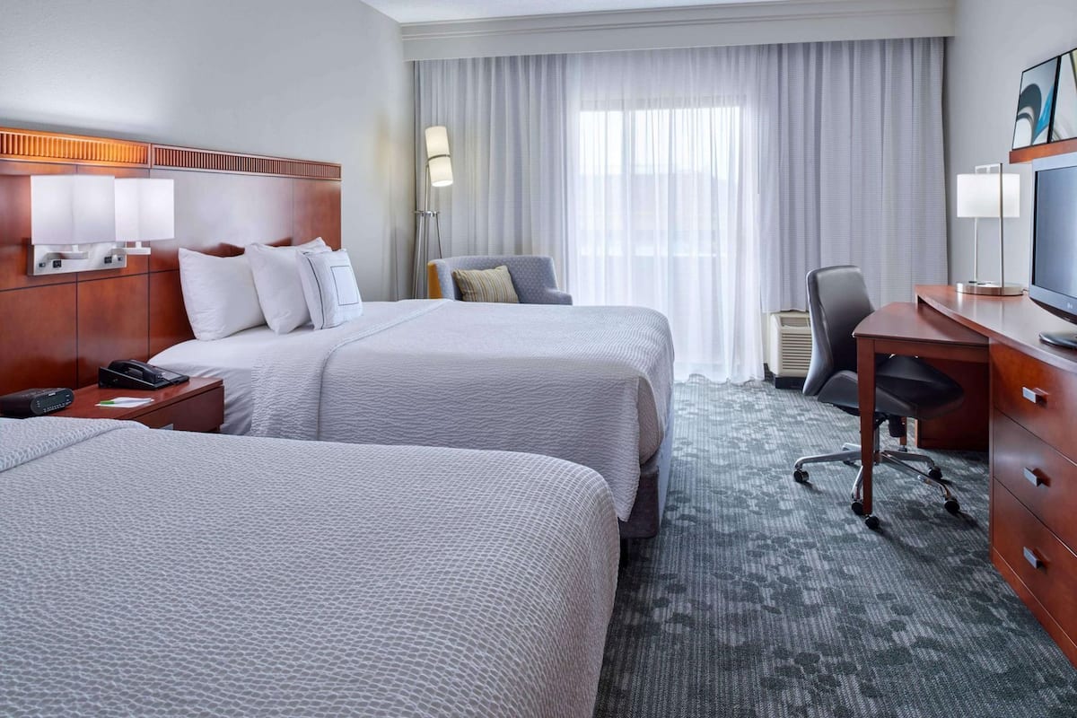 A hotel room with two beds, a desk, a chair, a TV, and a lamp.