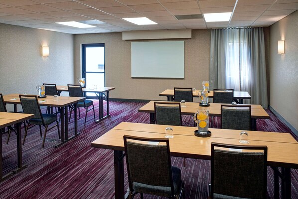 Un salón de conferencias con varias mesas y sillas, una pantalla de proyector y un ventanal con cortinas.