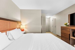 Una habitación de hotel con una cama grande, un cabecero de madera, una mesita de noche con una lámpara, un aparador de madera y un espejo.