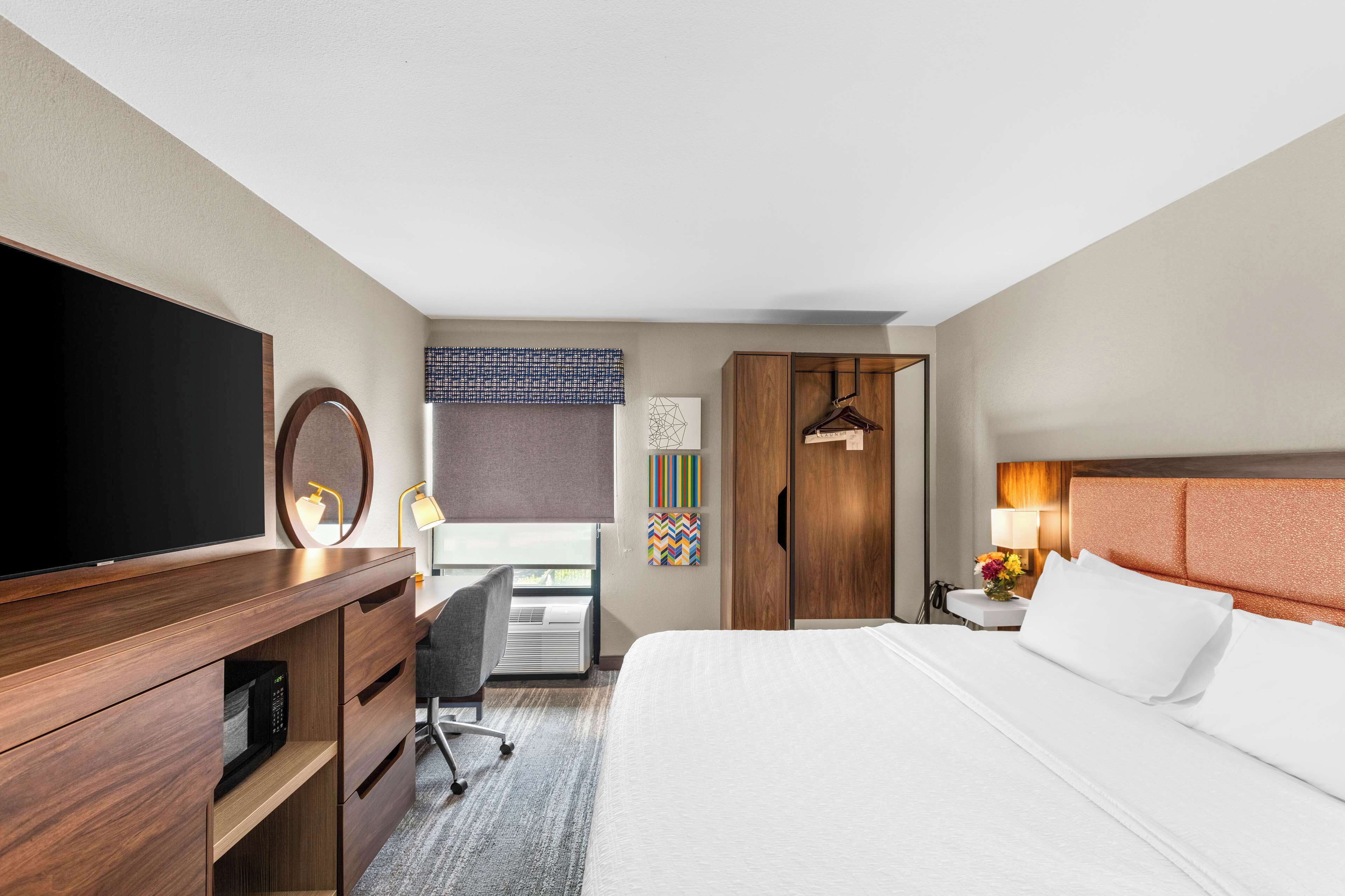 Habitación de hotel con cama, escritorio con silla, televisor, armario y ventana con persianas.