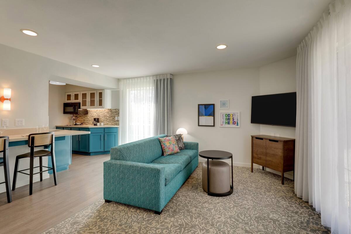 Una sala de estar moderna con un sofá color turquesa, una mesa de centro, un televisor y una zona de cocina con armarios azules y un salpicadero de piedra.
