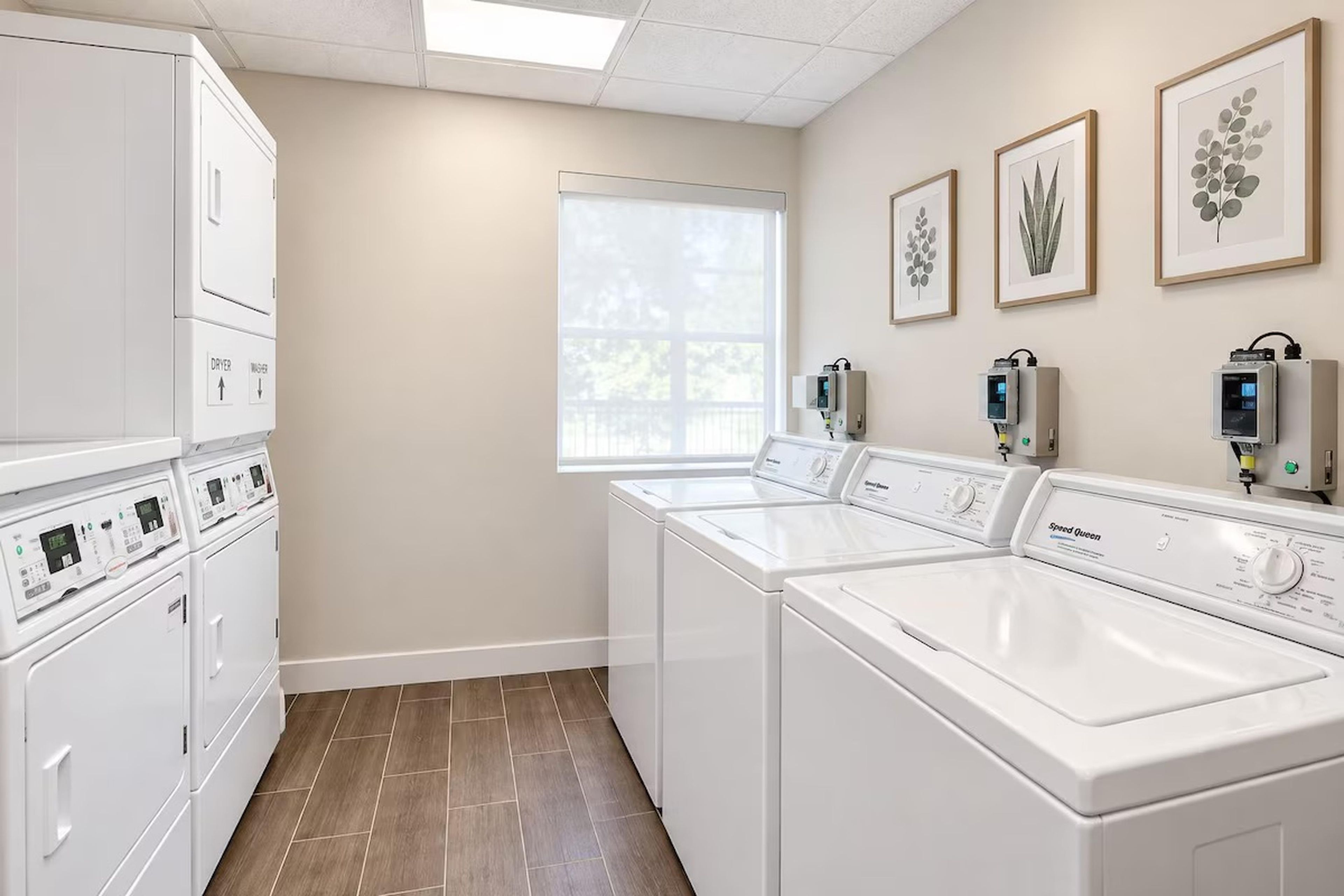 A laundry room with stacked washers and dryers, a window, and framed botanical prints on the wall.