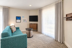 Habitación de hotel con un sofá color turquesa, una mesita redonda pequeña, un televisor en la pared y un armario de madera.
