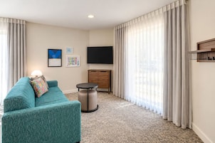 A hotel room with a teal sofa, a small round table, a TV on the wall, and a wooden cabinet.