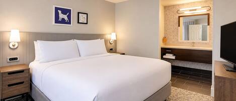 A hotel room with a large bed, two bedside tables, a TV, a bathroom with a mirror, and artwork on the wall.