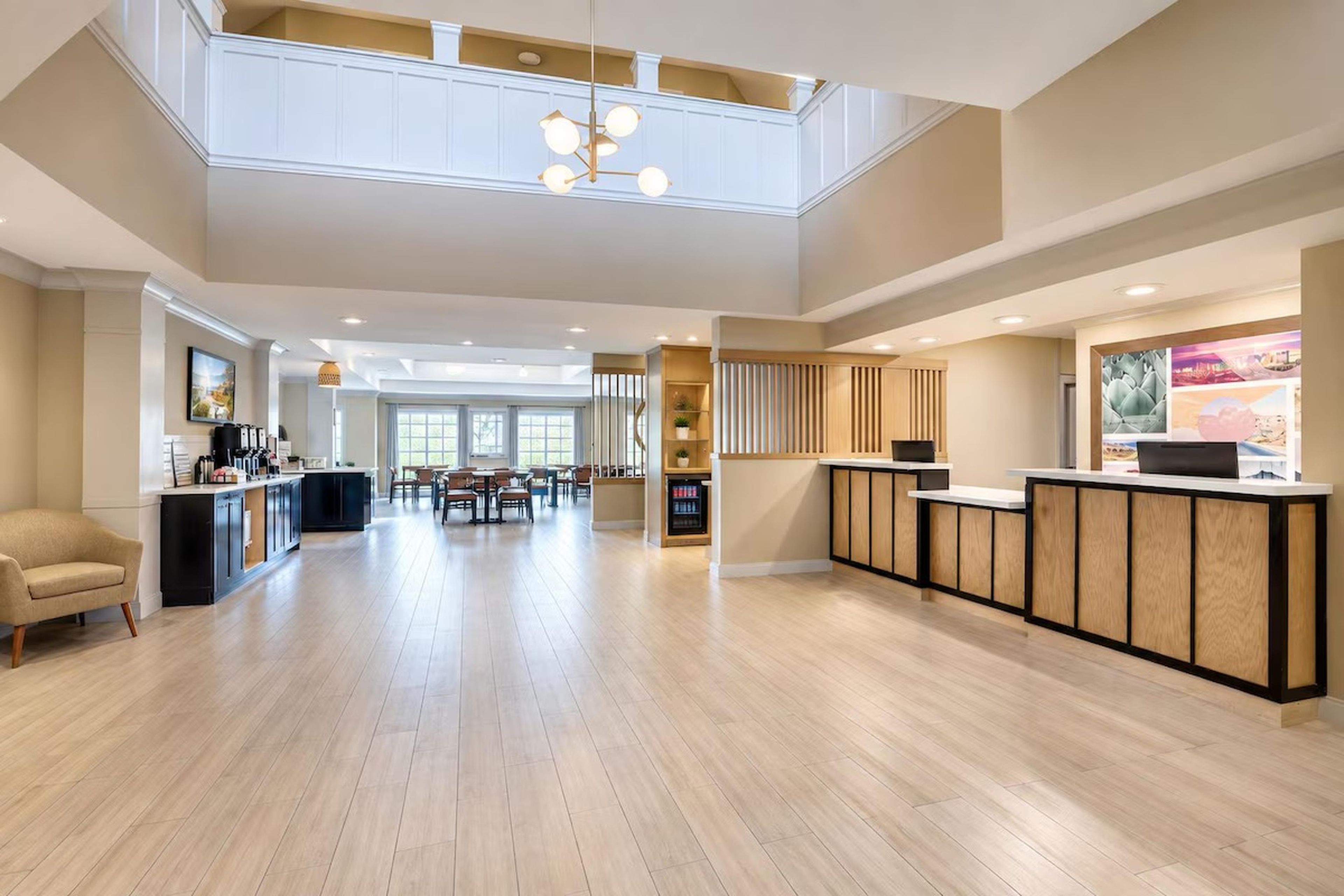 A spacious hotel lobby with a high ceiling, a reception desk, a dining area, and a kitchen visible in the background.