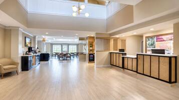 A spacious hotel lobby with a high ceiling, a reception desk, a dining area, and a kitchen visible in the background.