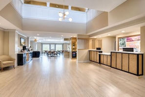 Un hall d’hôtel spacieux avec un plafond haut, un comptoir d’accueil, un espace repas et une cuisine visible au fond.