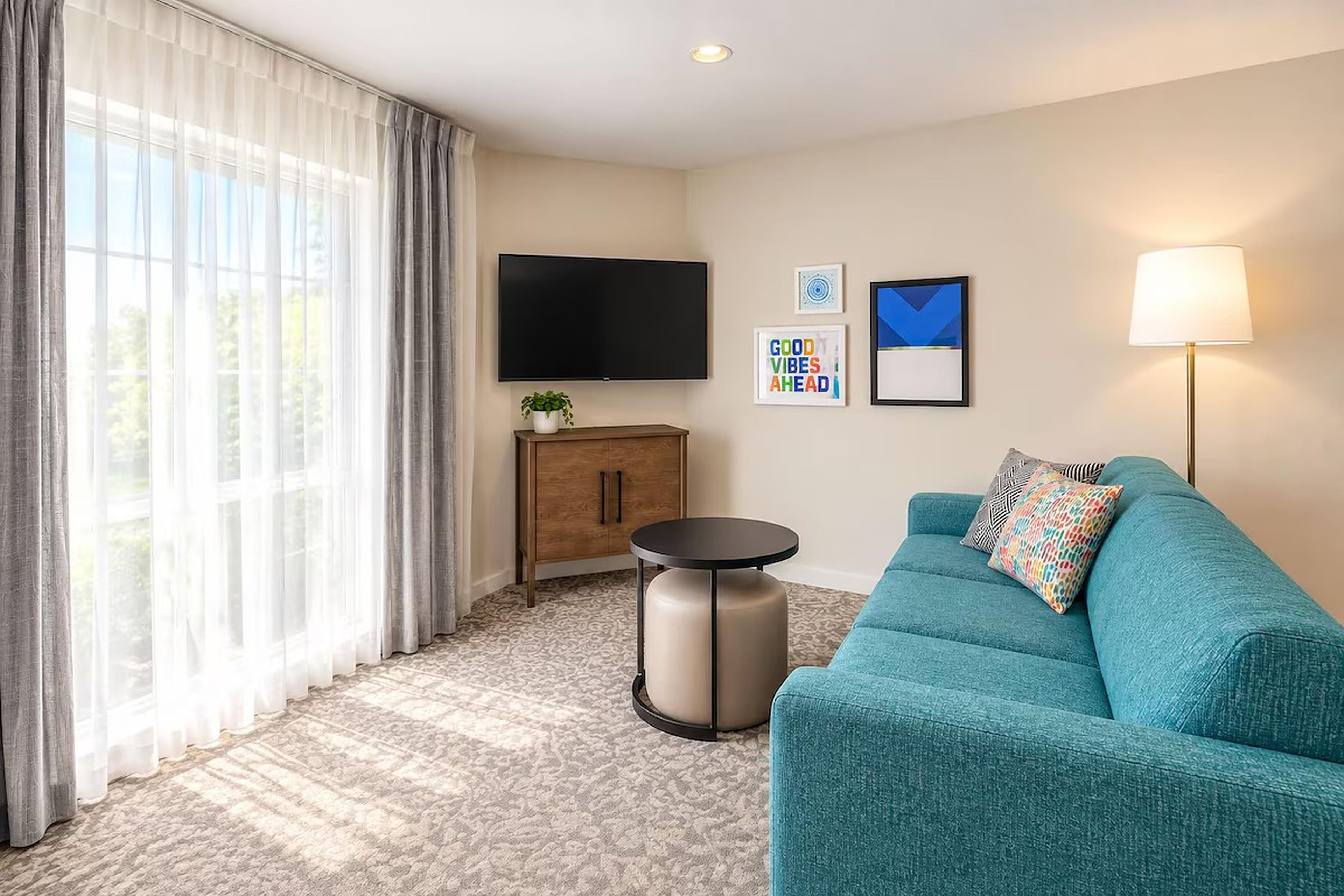 Una sala moderna con un sofá azul turquesa, un televisor de pantalla plana, un mueble aparador, una mesita redonda y una lámpara.