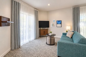 Habitación de hotel con sofá, televisor, una mesita con una planta y un mueble de televisión montado en la pared.