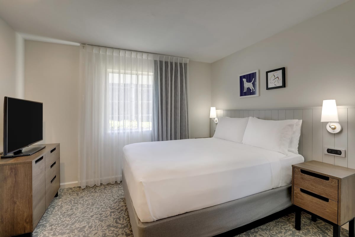 Habitación de hotel con cama, televisor sobre una cómoda de madera, mesita de noche con lámpara y dos lámparas de pared.