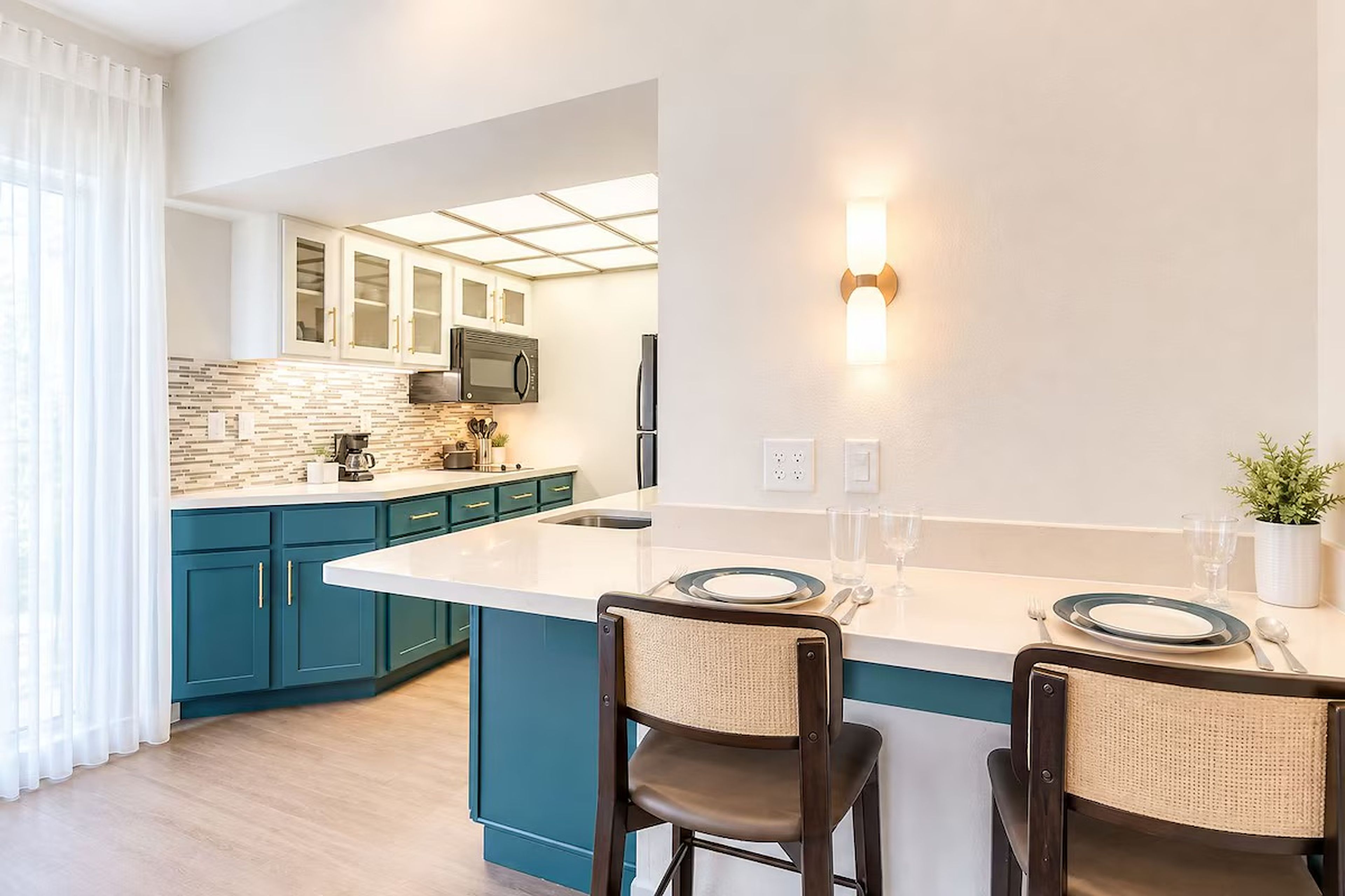 A modern kitchen with teal cabinets, a white countertop, and a built-in microwave. There are two bar stools with woven seats.