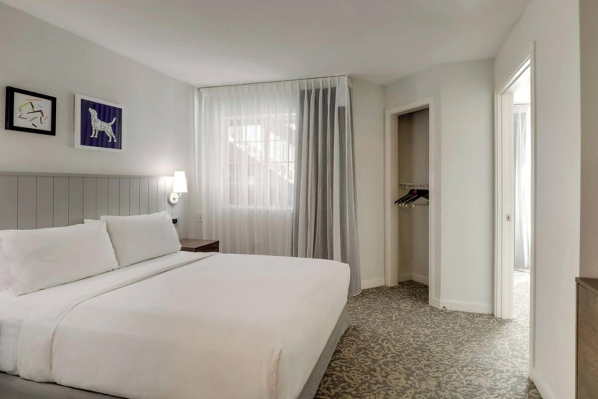 Una habitación de hotel con una cama grande, una mesita de noche con lámpara, un armario y una puerta que da a otra habitación.