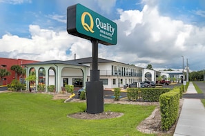 A hotel sign with a logo and name, a building with a green roof and arched windows, and a parking area with cars.