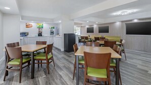A modern dining area with wooden tables and chairs, a kitchen area with white cabinets, and a large flat-screen TV mounted on the wall.