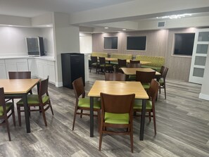 Een moderne eetruimte met houten tafels en stoelen, een keuken met witte kasten, en een grote flatscreen aan de muur.
