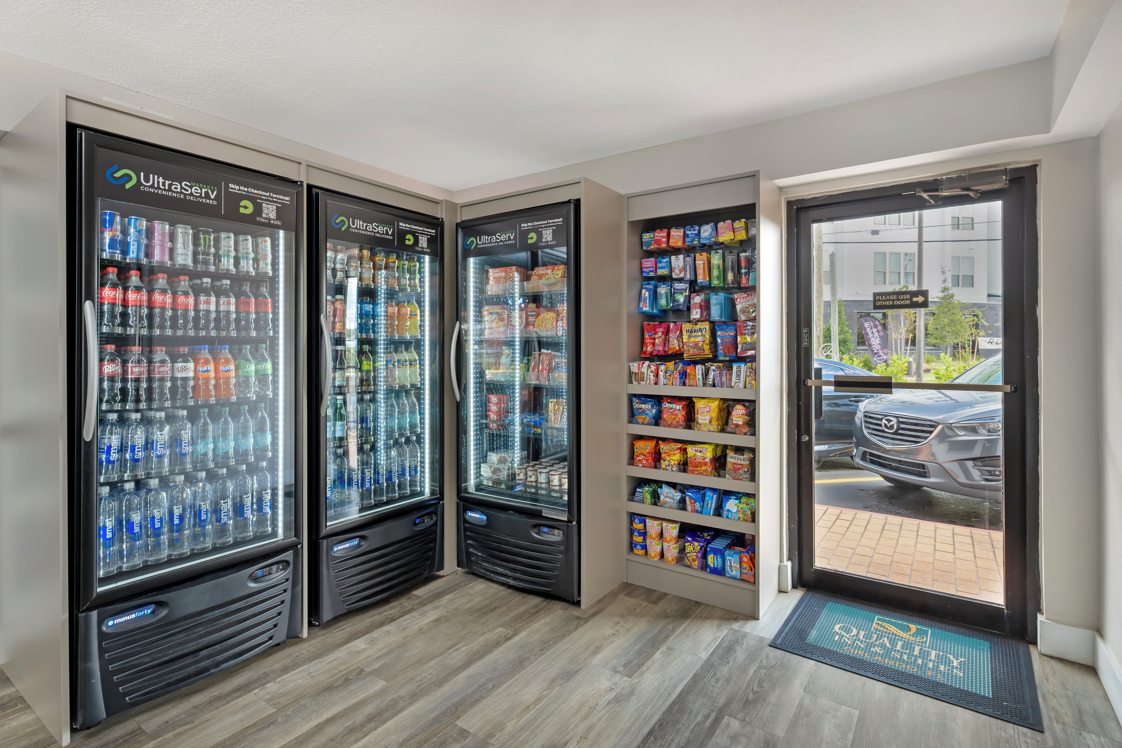 Une rangée de distributeurs automatiques de boissons proposant une variété de boissons, une étagère remplie de chips et d’en-cas, et une vue sur une voiture garée à l’extérieur.
