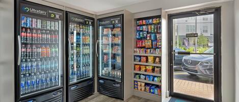 Una hilera de máquinas expendedoras de bebidas con diversas opciones, un estante con papas fritas y otros refrigerios, y una vista de un coche estacionado afuera.