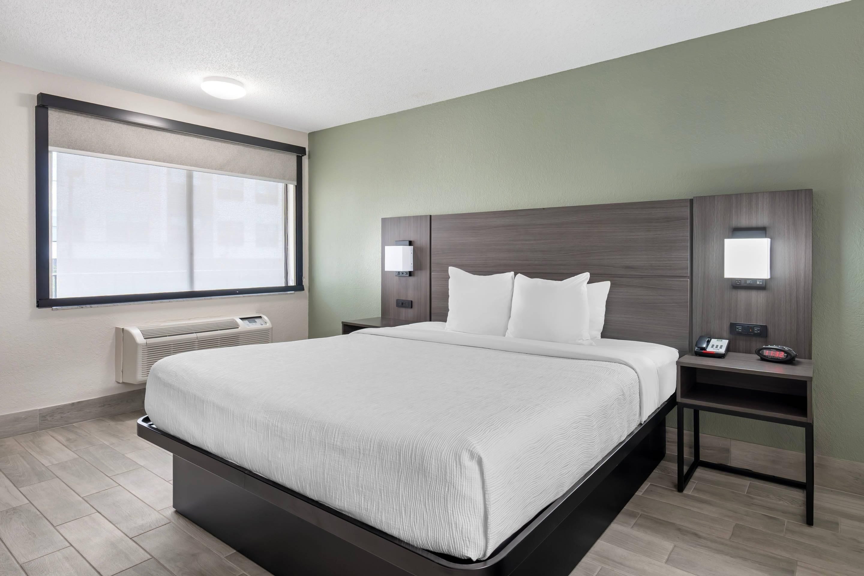 Quarto de hotel com uma cama grande, mesa de cabeceira com telefone e despertador, janela com persianas e aquecedor fixado na parede.
