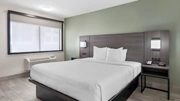 Quarto de hotel com uma cama grande, mesa de cabeceira com telefone e despertador, janela com persianas e aquecedor fixado na parede.