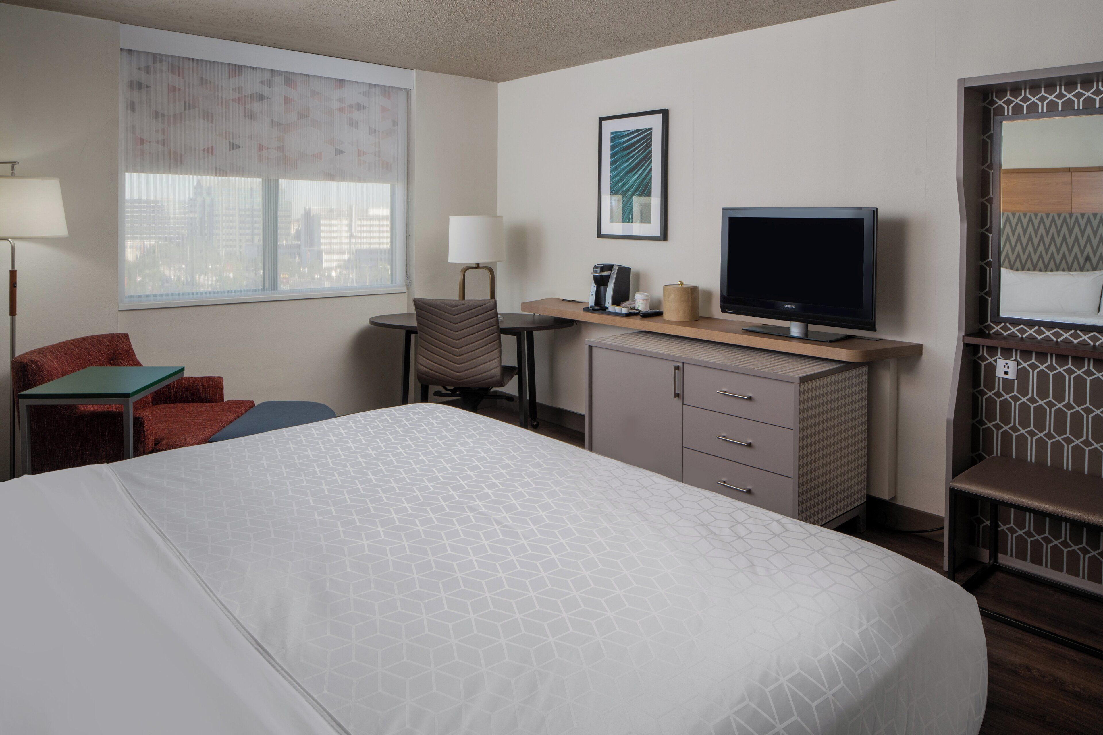 A hotel room with a bed, a desk with a television, a chair, a lamp, and a window with a city view.