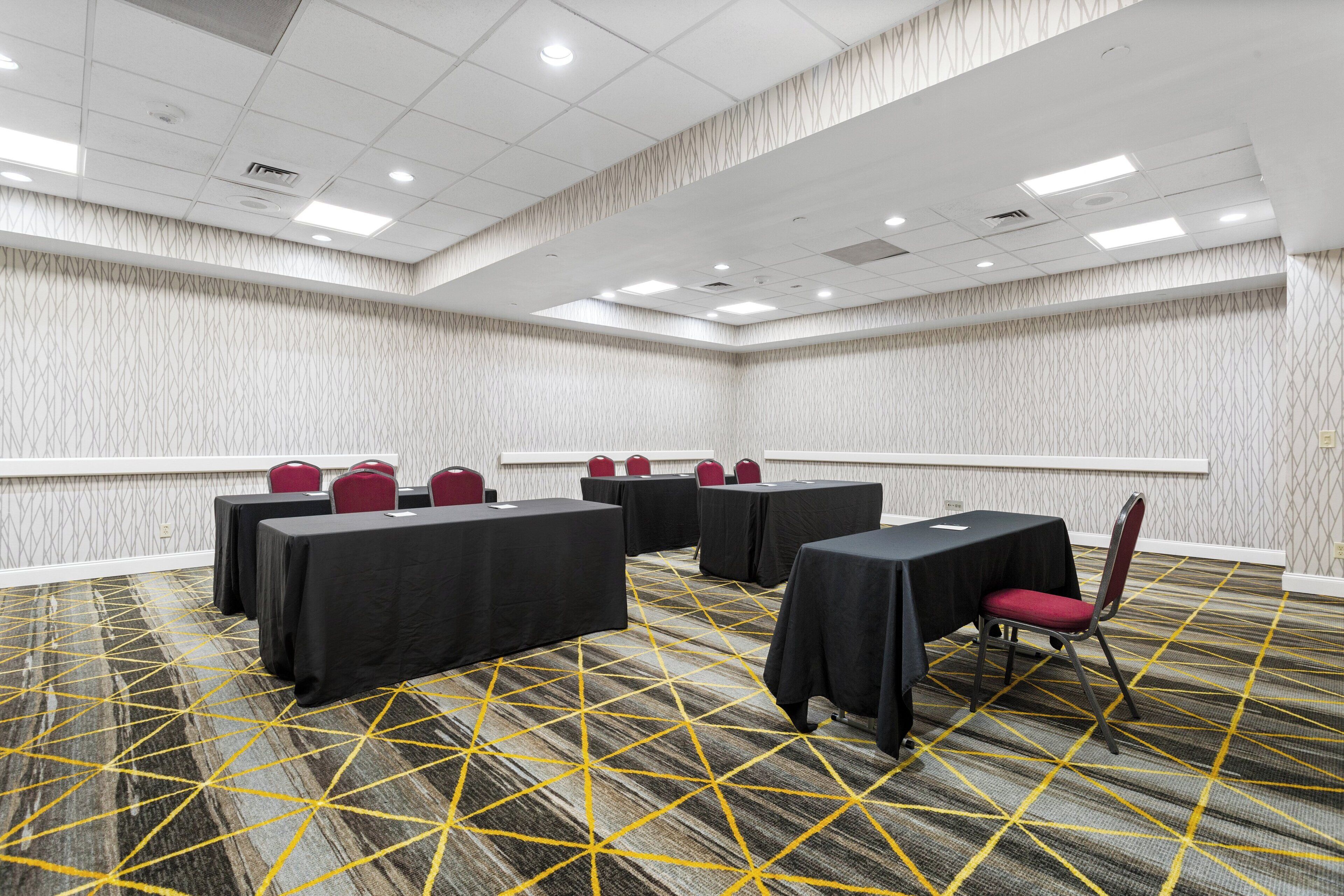 A conference room with tables and chairs, patterned carpet, and a high ceiling with recessed lighting.