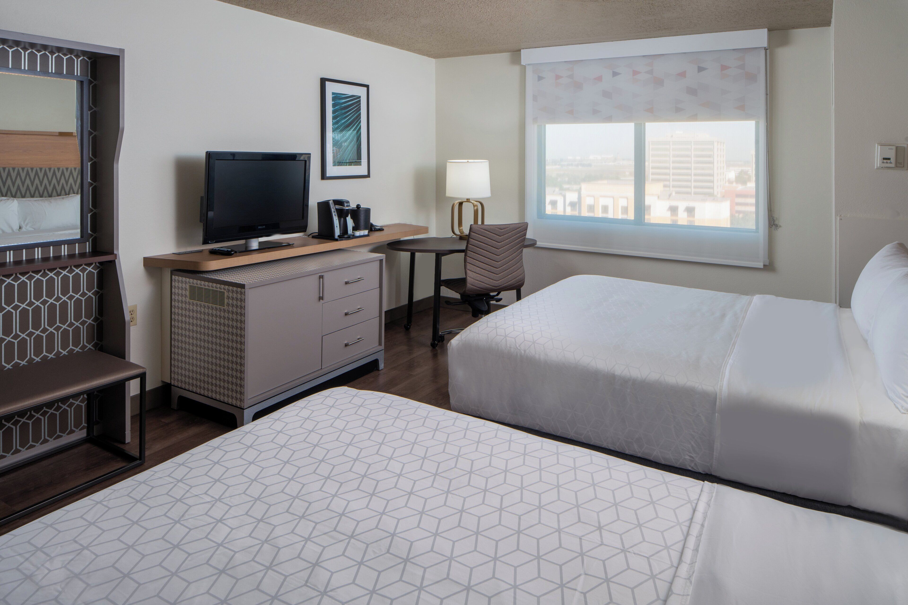 A hotel room with two beds, a desk with a lamp, a television, and a window with a view of buildings.