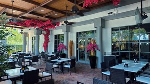 Un espacio exterior para comer con mesas y sillas, una entrada al bar y plantas decorativas.