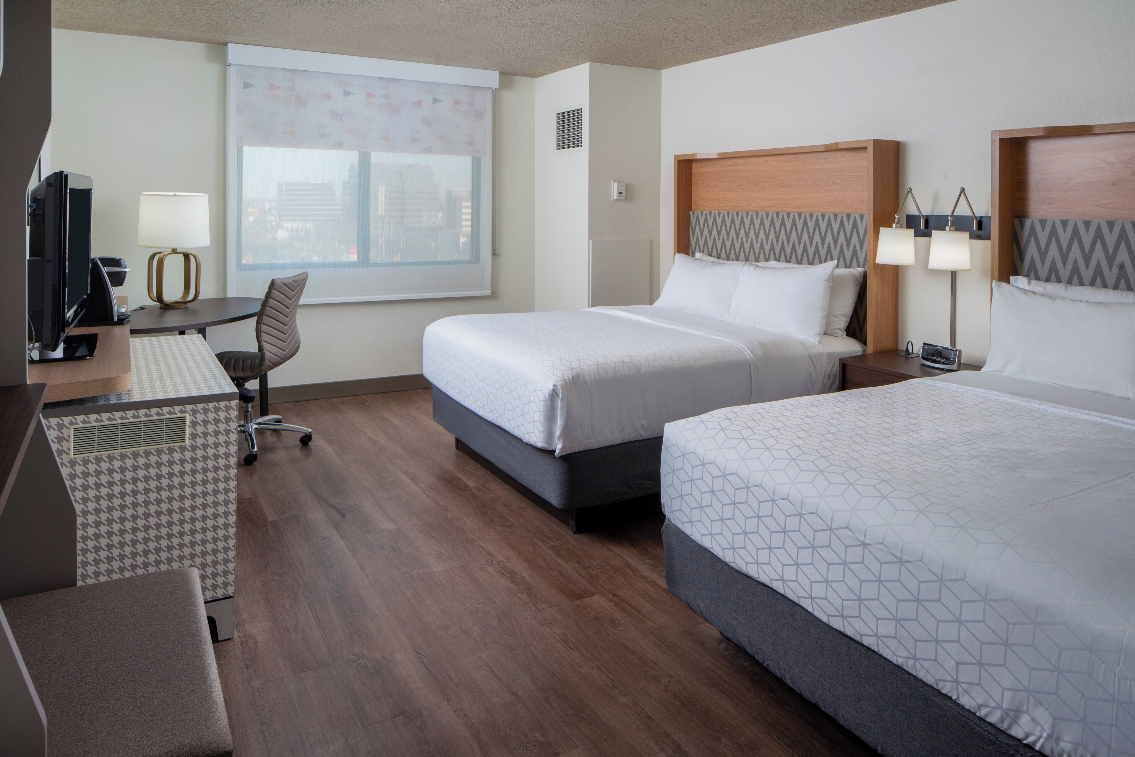 A hotel room with two beds, a desk with a computer, a chair, and a window with blinds.