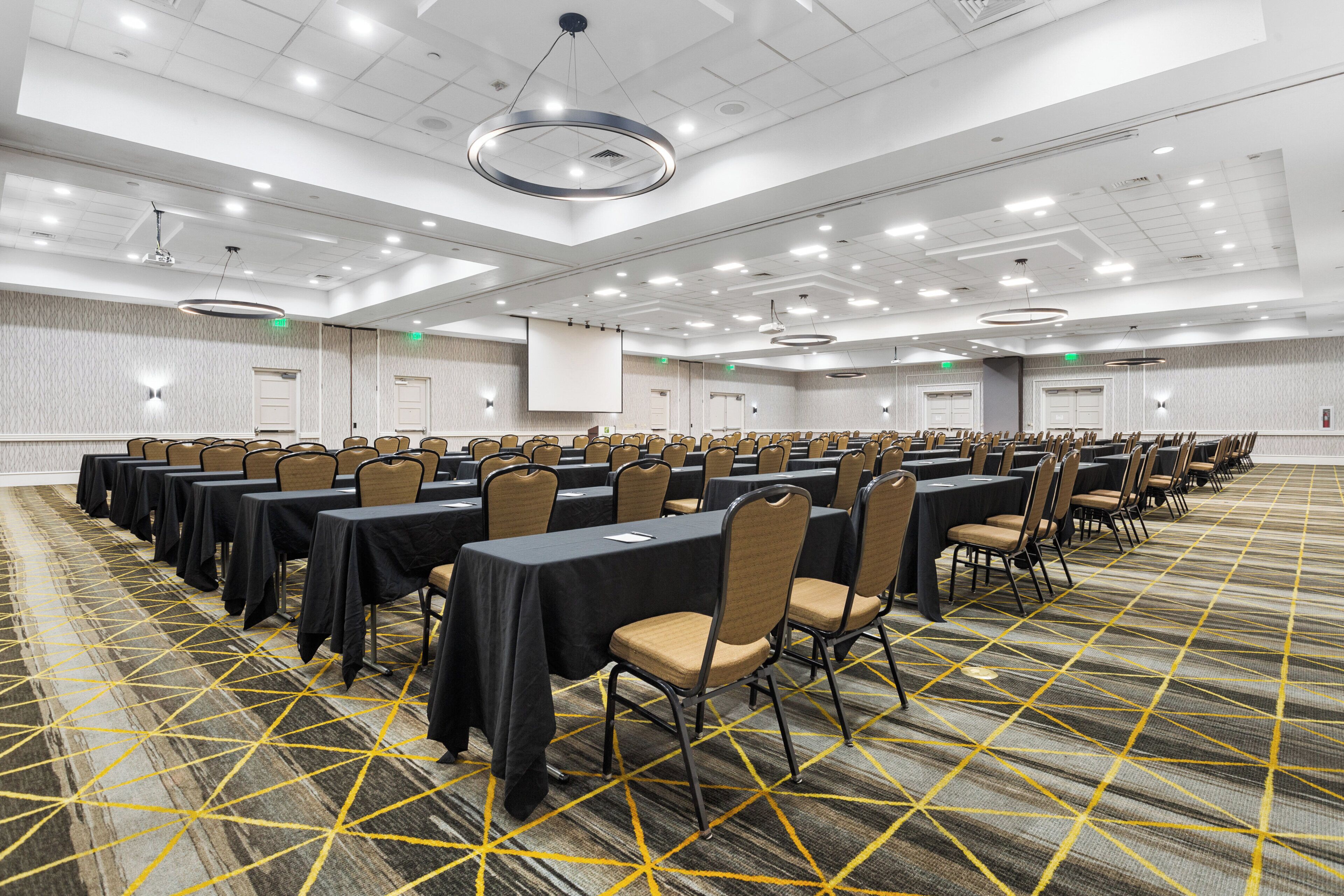 A large conference room with rows of tables and chairs, a carpeted floor, and a projector screen.