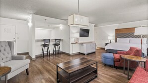 Una habitación de hotel moderna con una cama, un sillón gris, una mesa de centro de madera, un televisor y una kitchenette.