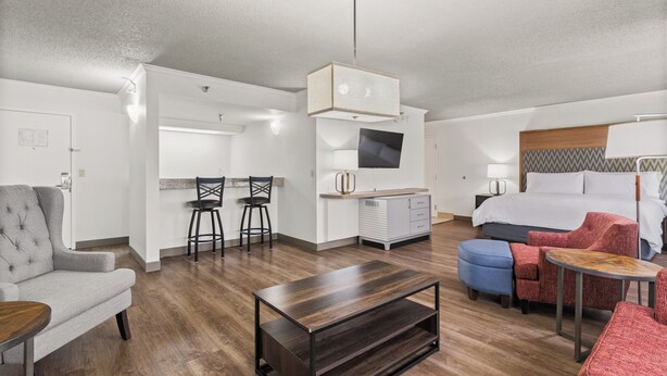 Una habitación de hotel moderna con una cama, un sillón gris, una mesa de centro de madera, un televisor y una kitchenette.