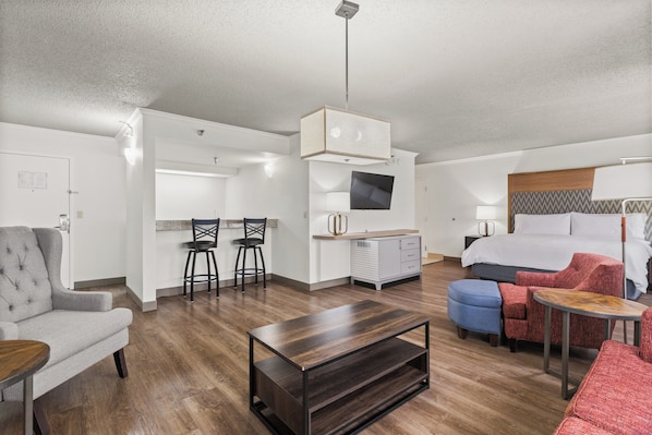Una habitación de hotel moderna con una cama, un sillón gris, una mesa de centro de madera, un televisor y una kitchenette.