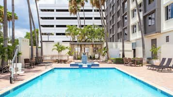Una piscina con fuente acuática, rodeada de sillas y mesas de descanso, con un edificio moderno al fondo.