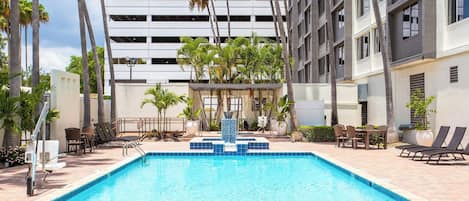 Una piscina con fuente acuática, rodeada de sillas y mesas de descanso, con un edificio moderno al fondo.