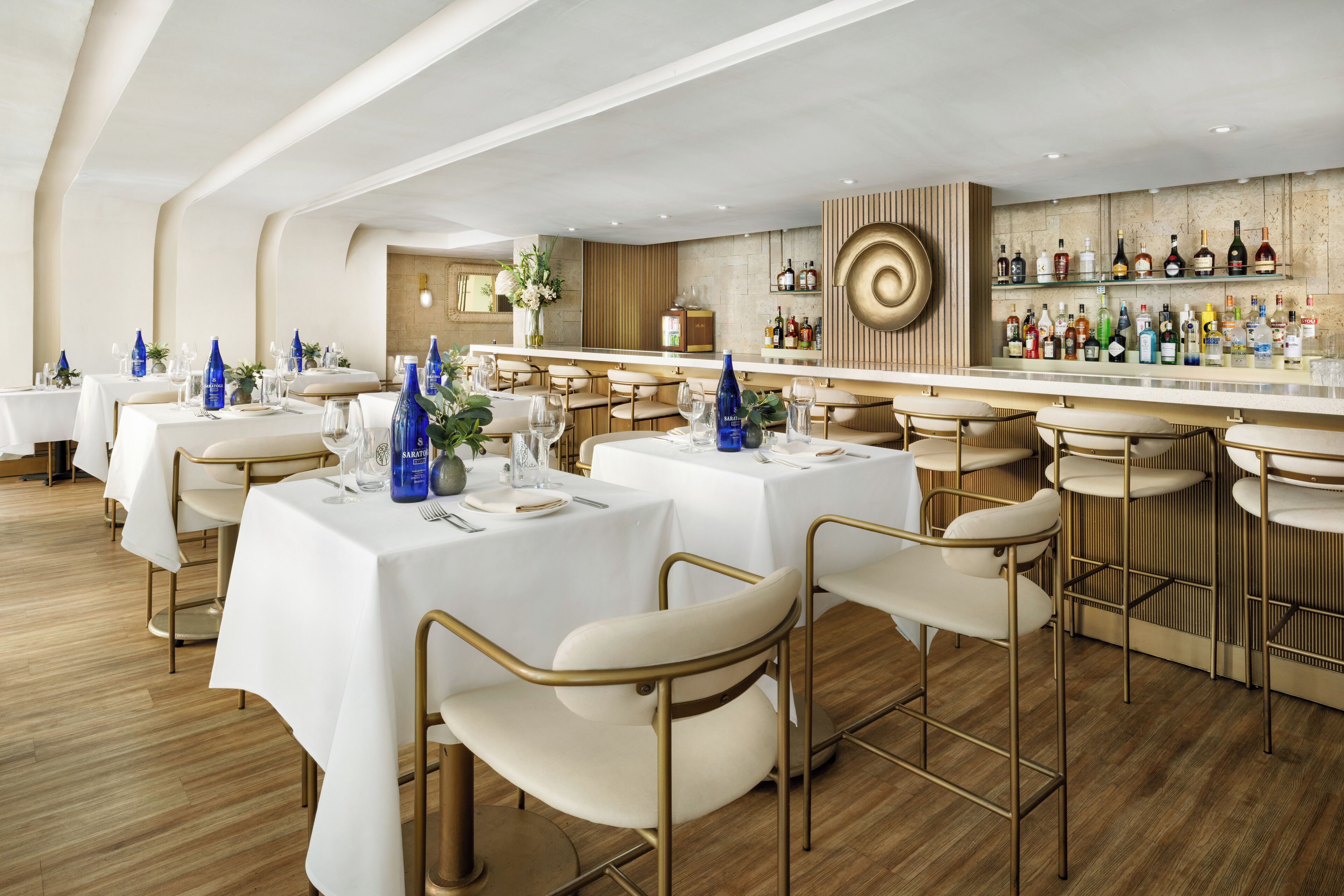 Un restaurant moderne avec des nappes blanches, des chaises dorées et un bar où sont exposées différentes bouteilles de spiritueux.