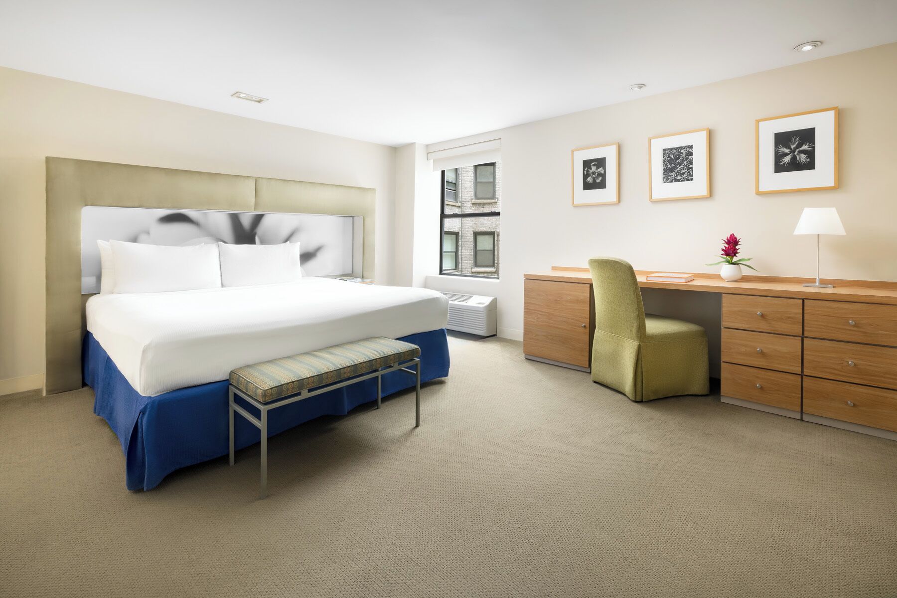 Hotelkamer met een groot bed, een bureau met stoel, een kastje en een raam met uitzicht op gebouwen.