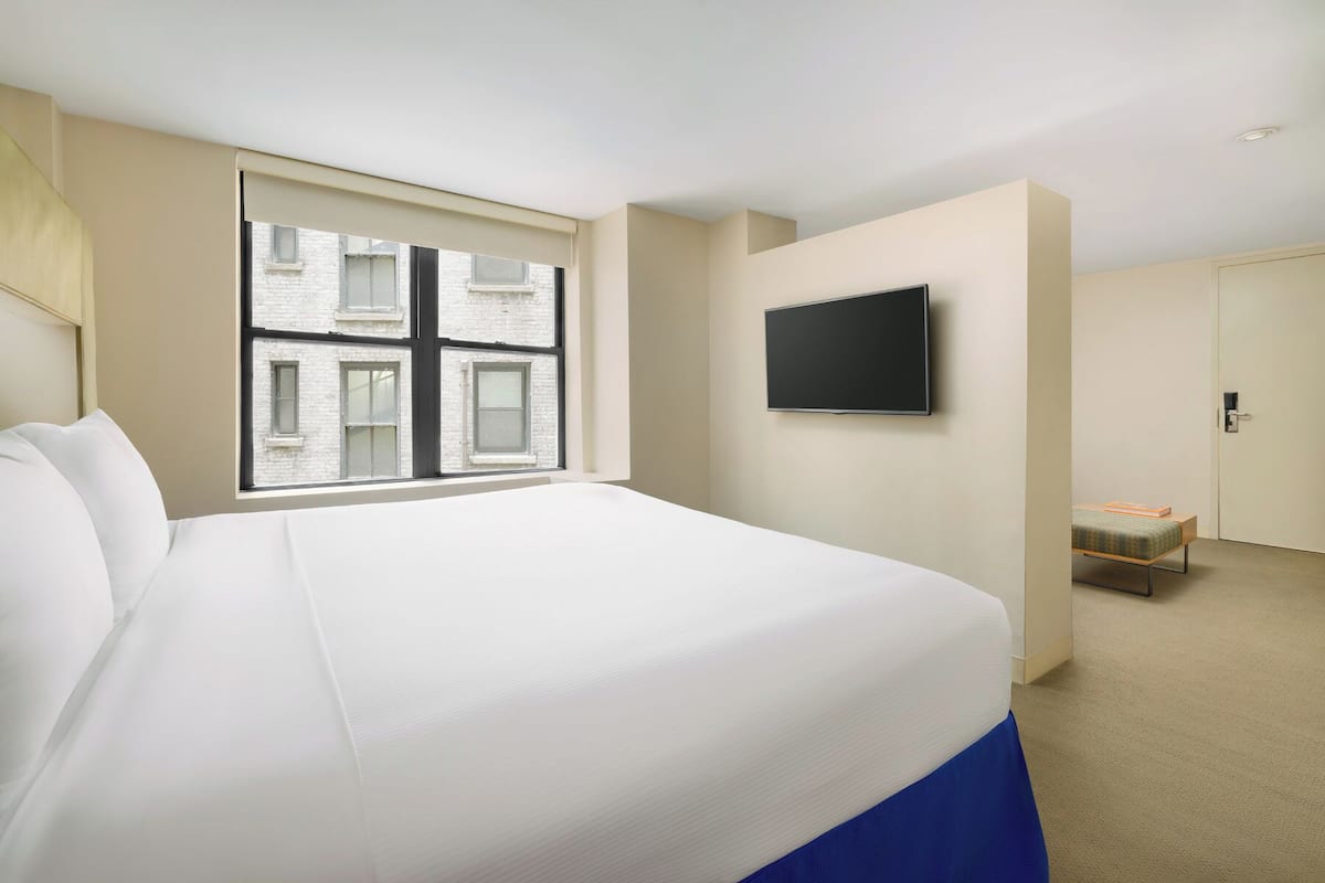A hotel room with a large bed, a TV mounted on the wall, and a window with a view of buildings.