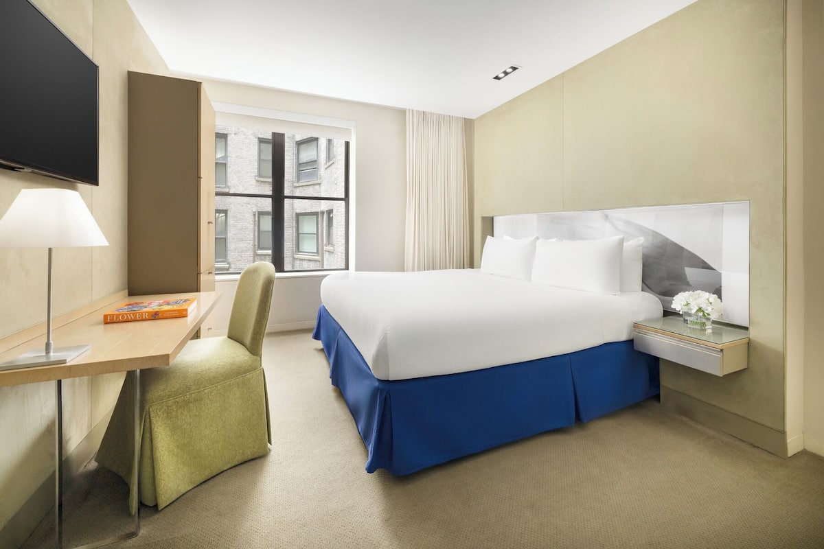 A hotel room with a bed, a desk, a chair, a TV, a window, and a nightstand with a vase of flowers.