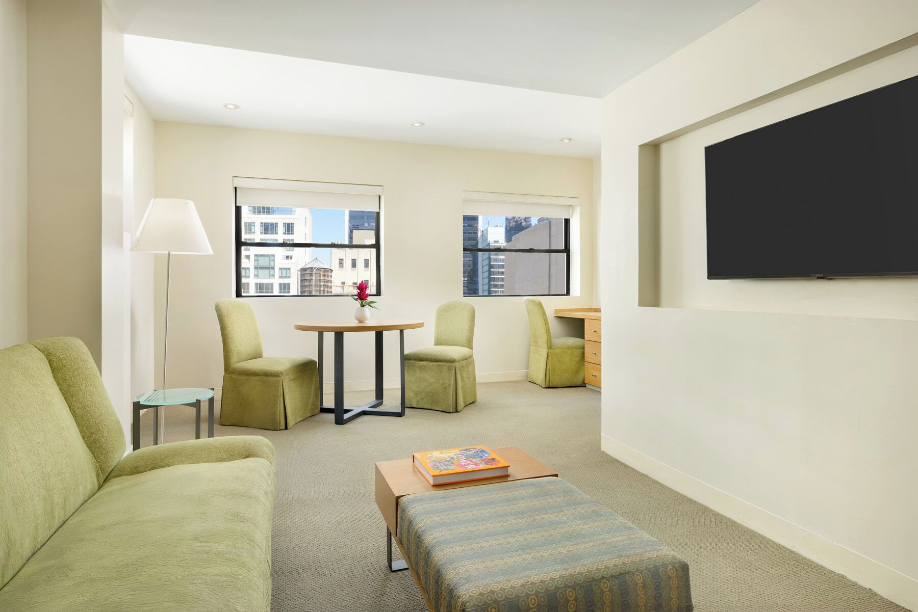 A modern hotel room with a sofa, two armchairs, a small table with a vase, a flat-screen TV mounted on the wall, and a window with a city view.