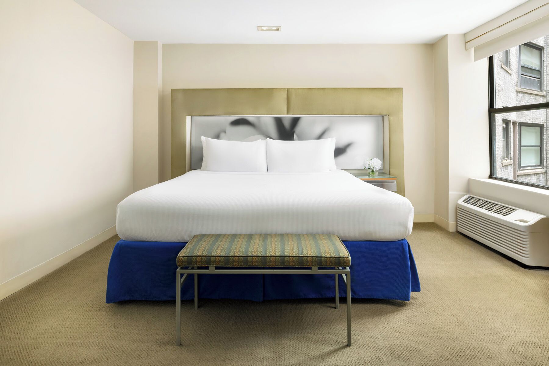 Une chambre d’hôtel avec un grand lit, une table de chevet, un banc et une fenêtre donnant sur des immeubles.