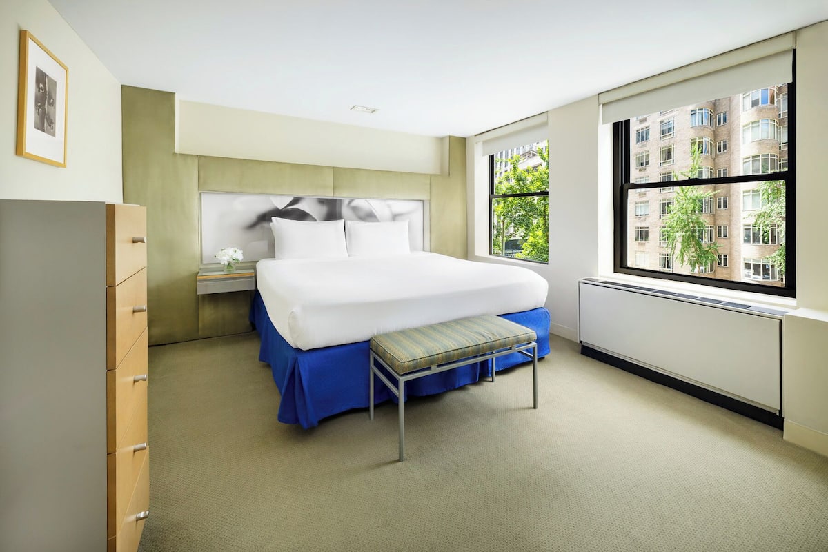 A modern hotel room with a large bed, a nightstand, a dresser, and a window with a city view.