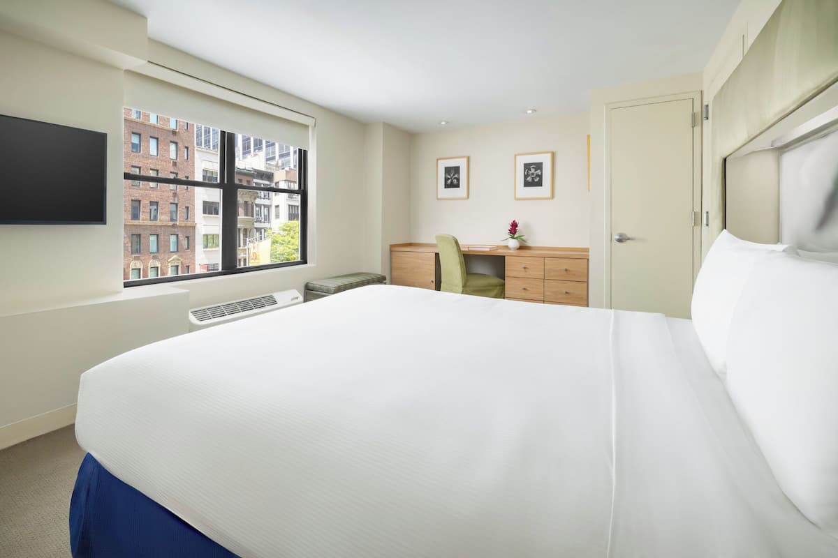 A hotel room with a large bed, a desk with a chair, a flat-screen TV mounted on the wall, and a window with a view of buildings.