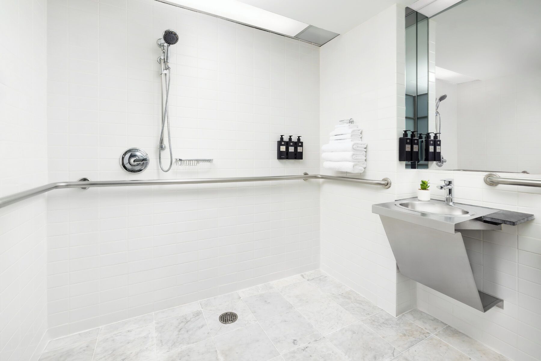 A modern bathroom with white tiled walls, a shower with a handheld hose, a grab bar, a sink with a mirror, and a small potted plant.