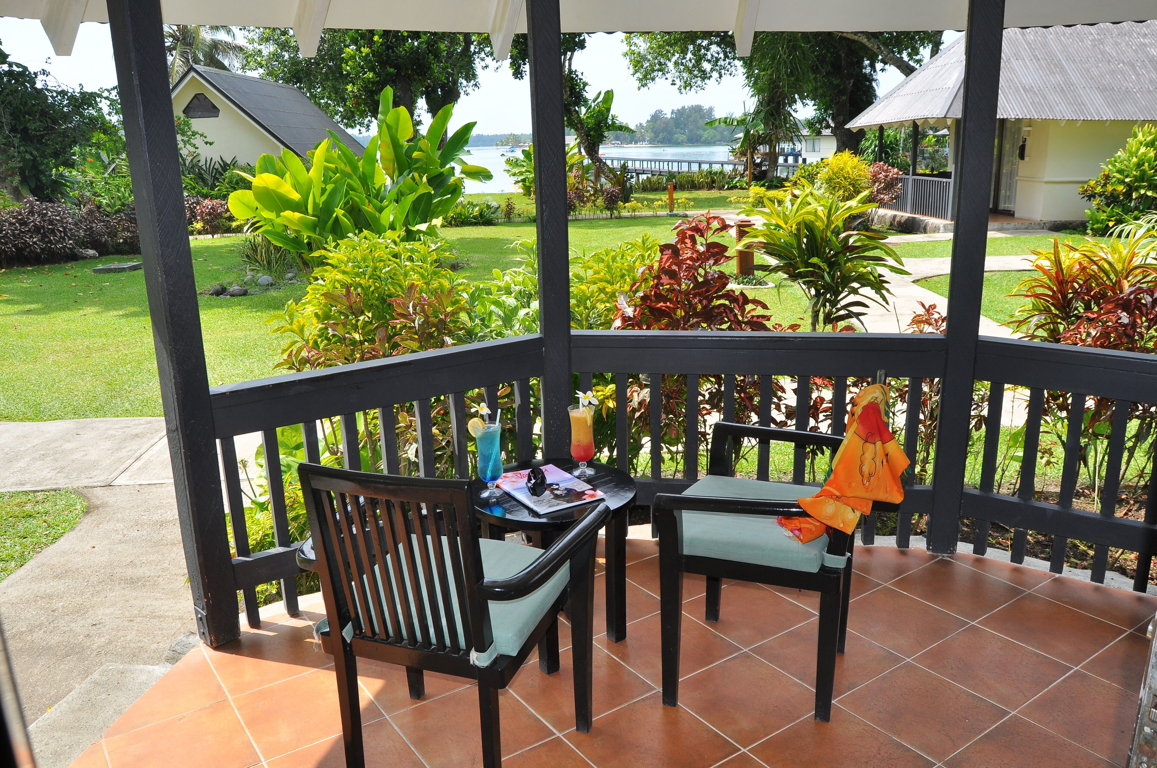 A covered outdoor seating area with two chairs and a table, overlooking a well-maintained garden and a body of water.