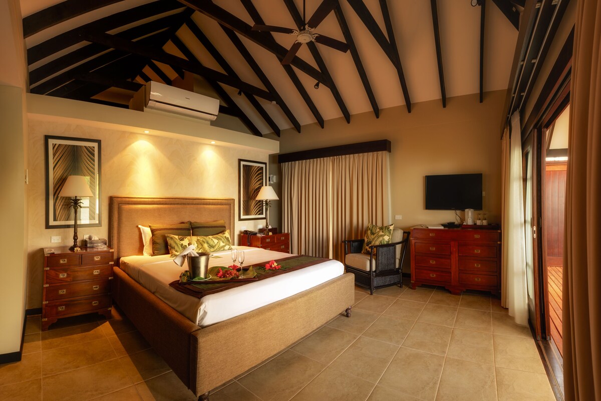 Een ruime slaapkamer met een groot bed, een flatscreen televisie, een houten dressoir en een plafondventilator.