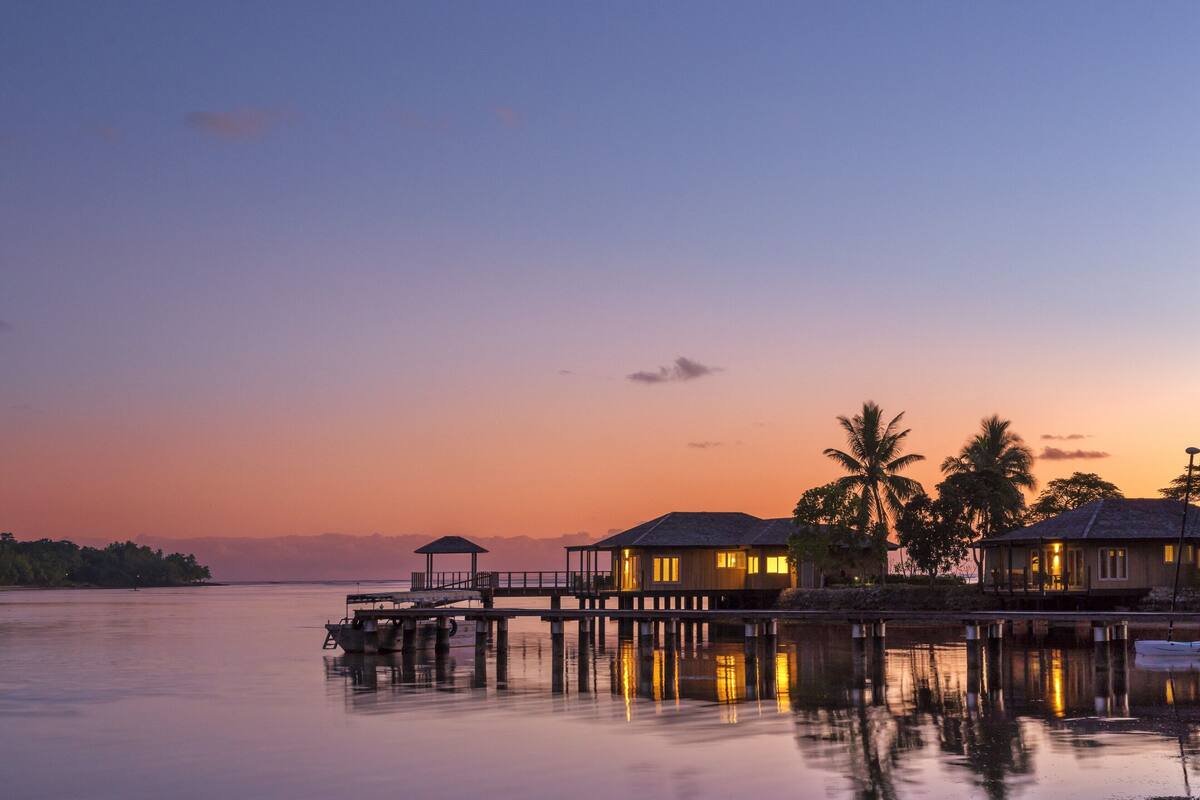 Ein Ufergrundstück mit Bungalows über dem Wasser, einer Mole und einem stillen See bei Sonnenuntergang.