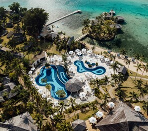 Vista aerea di un resort con una grande piscina, ombrelloni da spiaggia e palme.