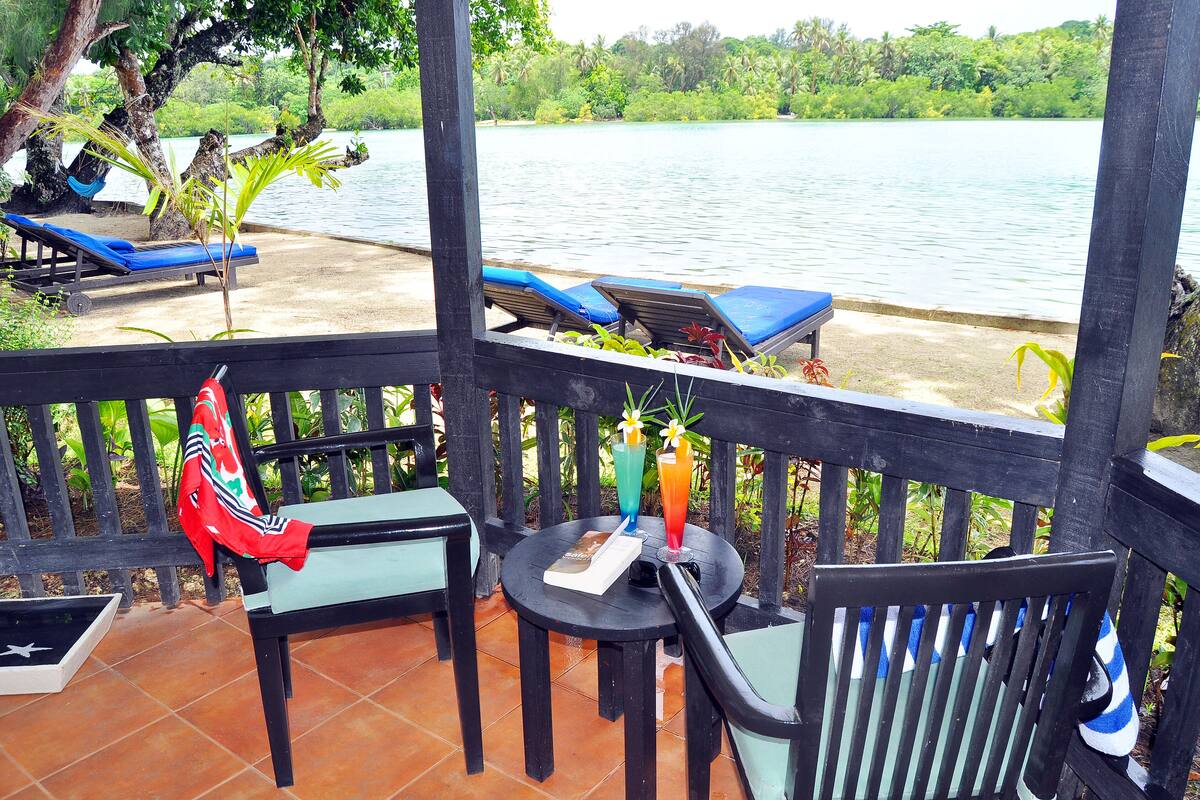 Eine Terrasse mit Seeblick, ausgestattet mit Liegestühlen, einem Tisch mit Getränken und einem Sessel mit rotem Kissen.